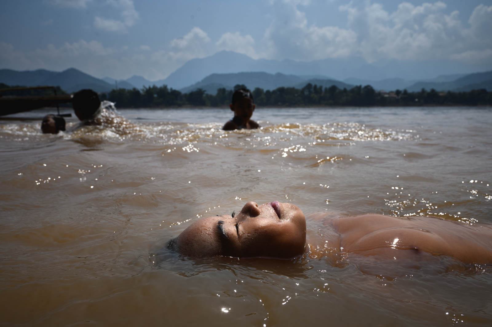 Ein laotisches Kind lässt sich auf dem Rücken durch den Mekong treiben, während weitere Kinder vor der Bergkulisse bei Luang Prabang baden.