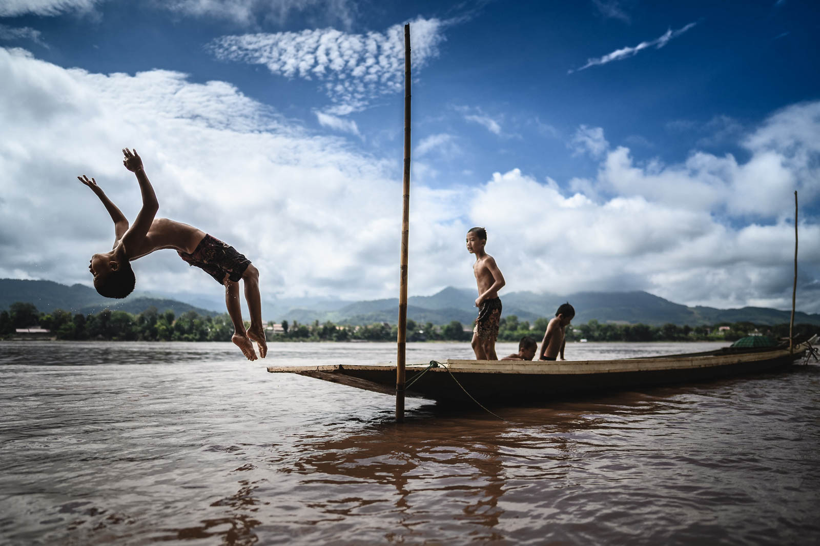 Laotische Kinder springen von einem schmalen Holzboot in den Mekong, umgeben von der Berglandschaft bei Luang Prabang.